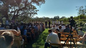 Wedding ceremony on the lawn during a sunny afternoon. Center aisle view from the back row of guests.